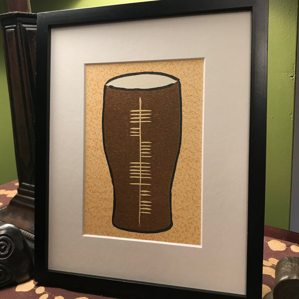 Painting of a pint glass with Ogham writing in the center, framed on a table.